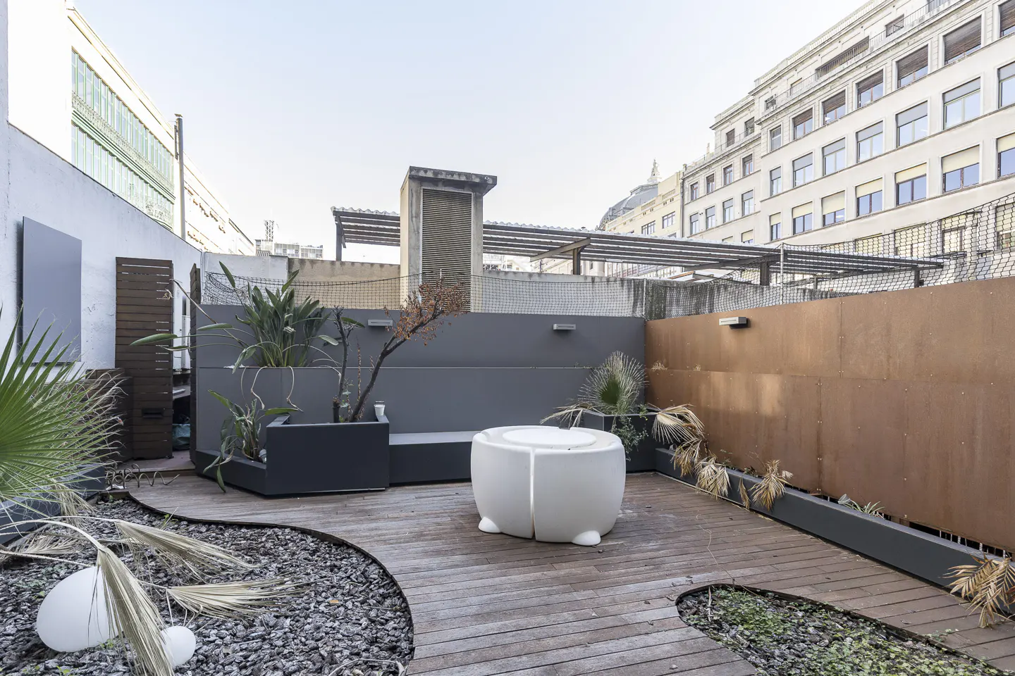 Modern rooftop terrace with wooden decking, a white circular table, potted plants, and a mix of grey and rusted metal walls.