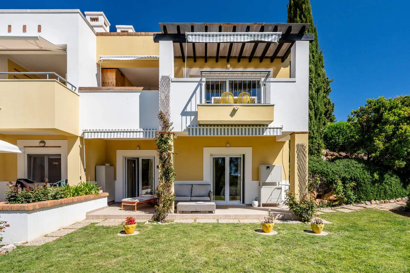 Exterior view of a two-story yellow and white building with a balcony, patio, and green lawn.