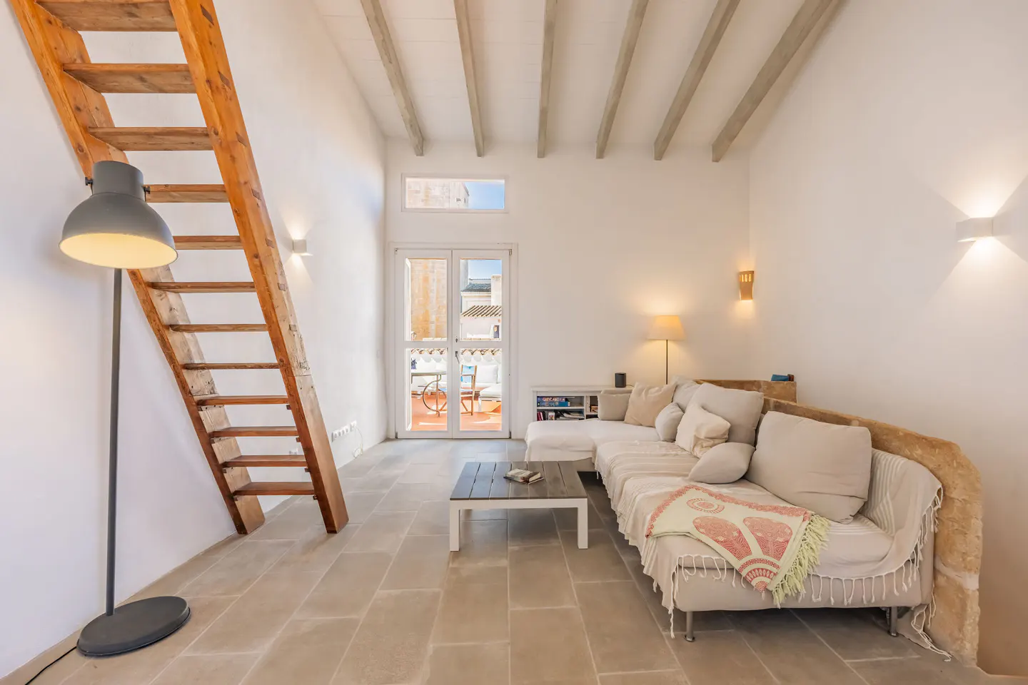 Bright living room with white walls, stone floors, and exposed beams. A wooden staircase leads up, and a large sofa faces a balcony.