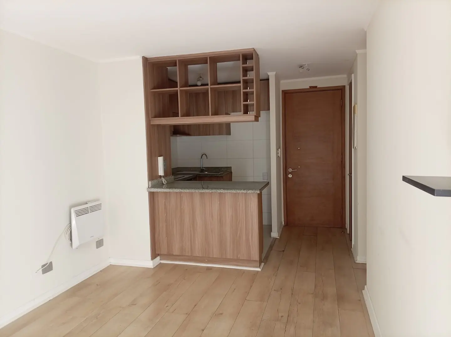 A bright apartment interior with wood floors, white walls, and a compact kitchen area with wood cabinets and a gray countertop. A brown door is visible in the background.