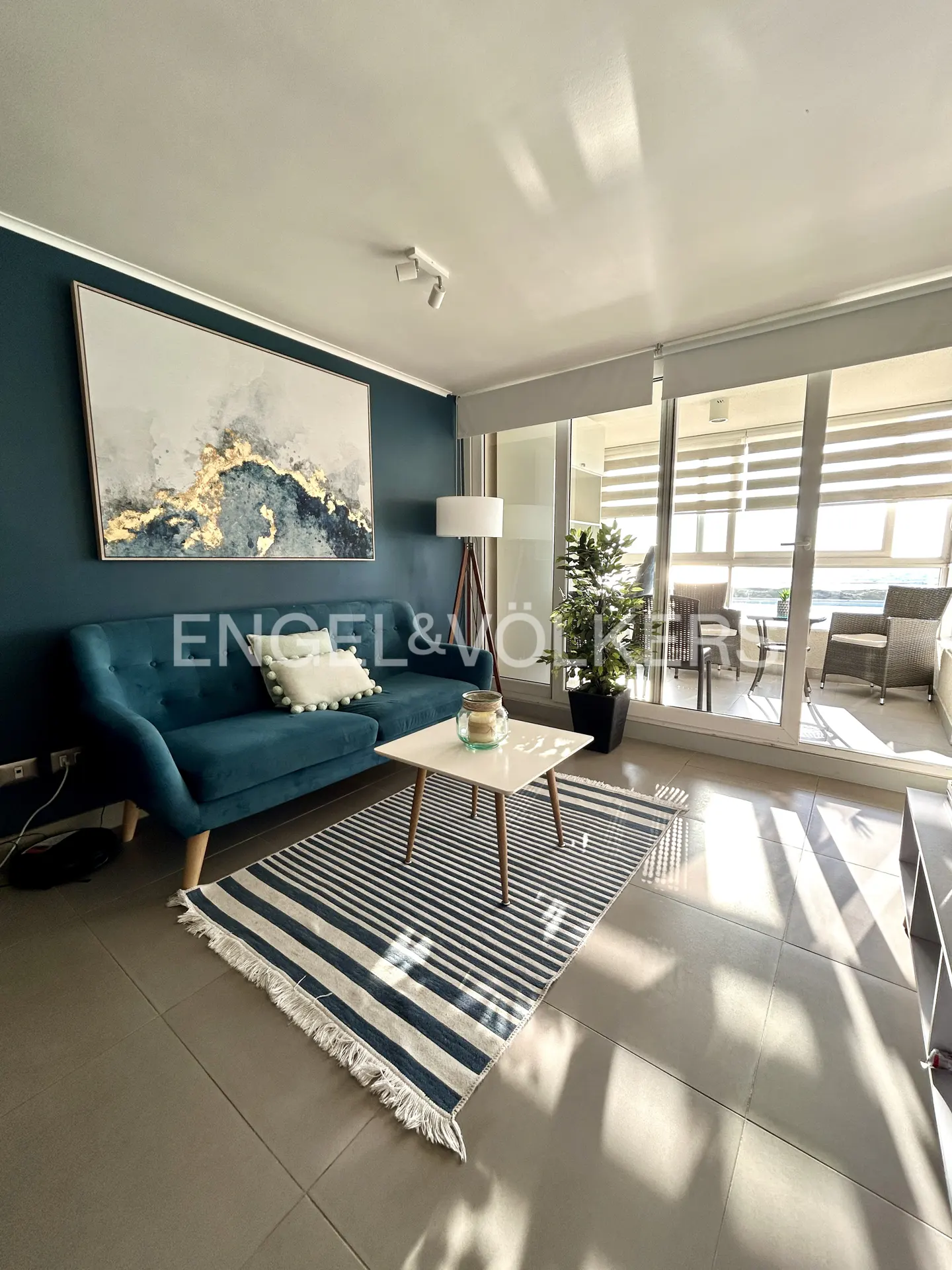 Modern living room with a teal sofa, abstract wall art, striped rug, and a balcony view through large glass doors.