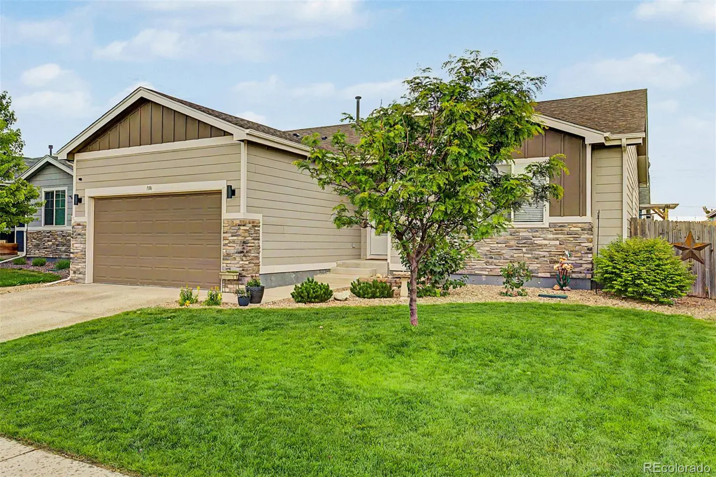 Single-story tan house with stone accents, a two-car garage, a green front lawn, and a small tree under a blue sky.