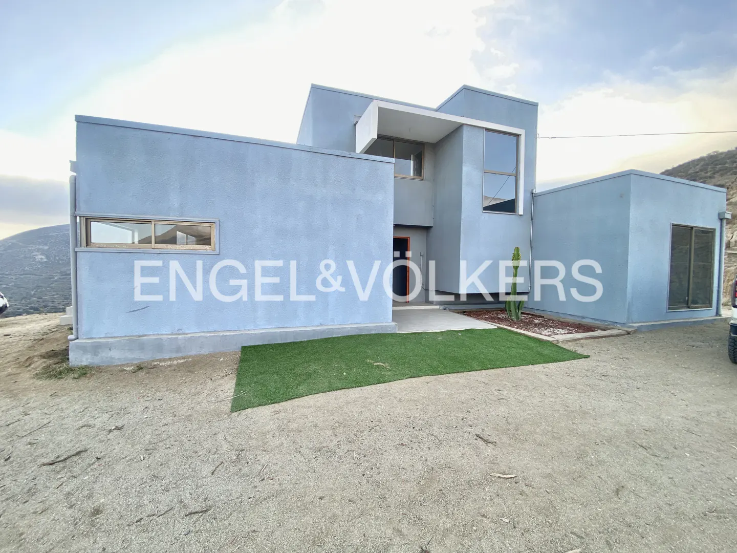 Modern blue house with geometric shapes, large windows, and a small patch of green grass in front. "Engel & Volkers" logo visible.