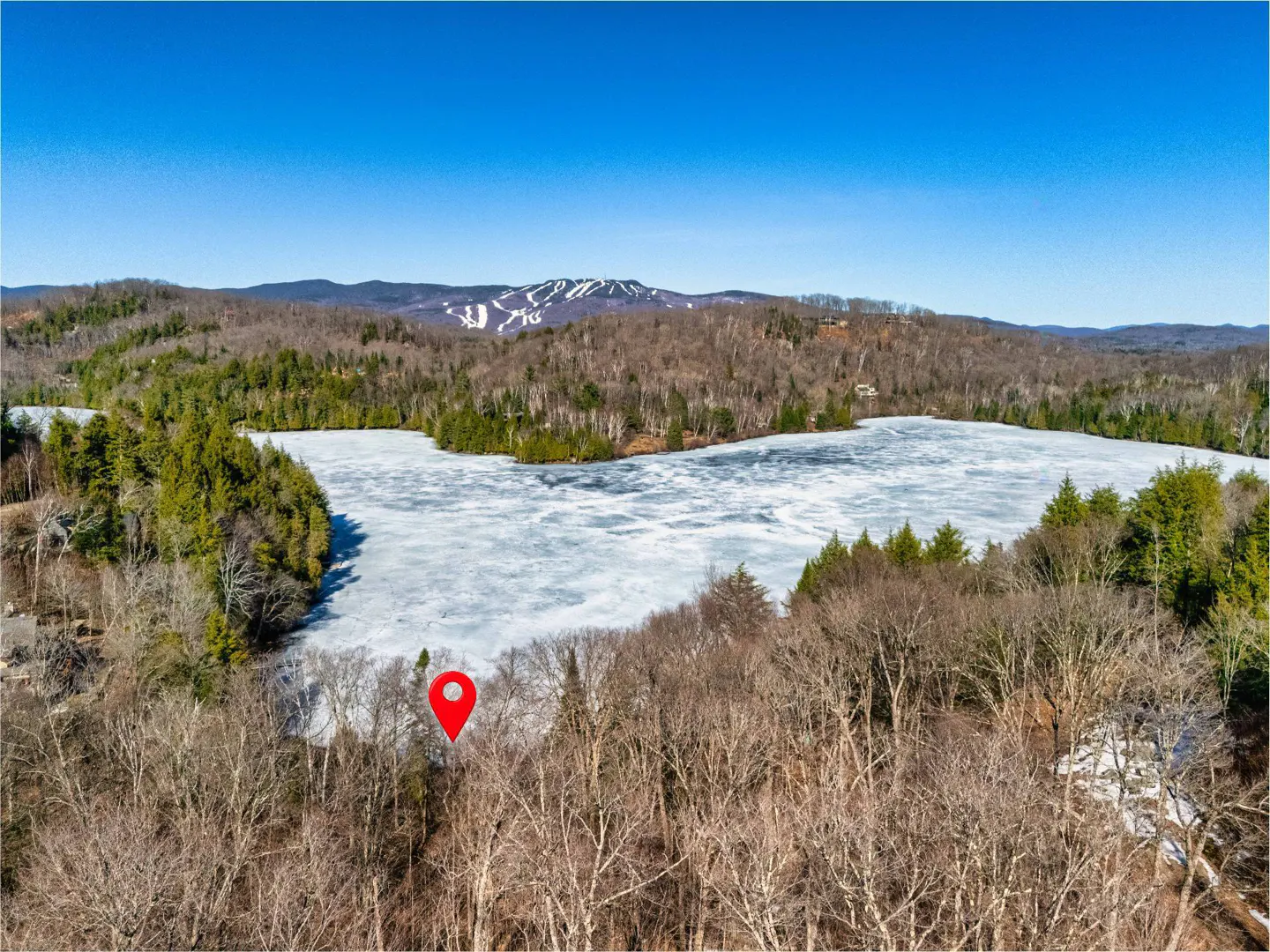 Aerial view of a partially frozen lake surrounded by trees, with a red pin marking a location near the shore. Ski slopes are visible on a distant mountain.