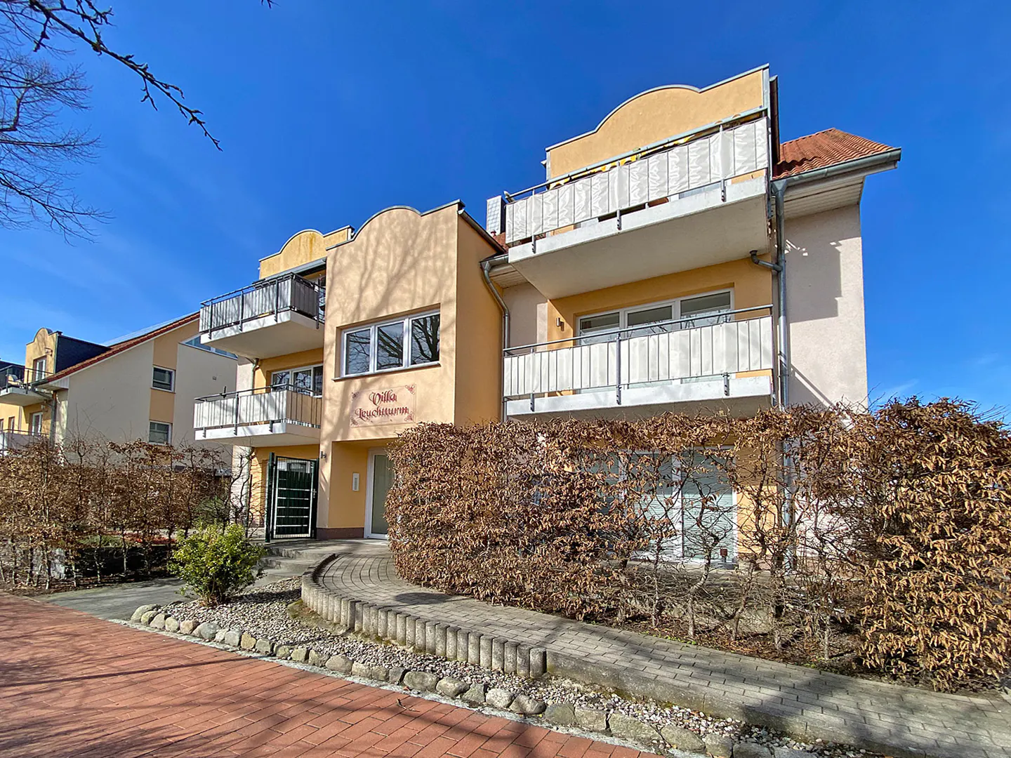Exterior of "Villa Leuchtturm," a yellow apartment building with balconies, hedges, and a brick walkway under a blue sky.
