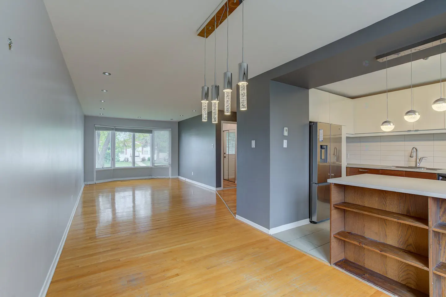 Open-concept living space with hardwood floors, gray walls, and modern lighting fixtures. Kitchen with stainless steel appliances visible.