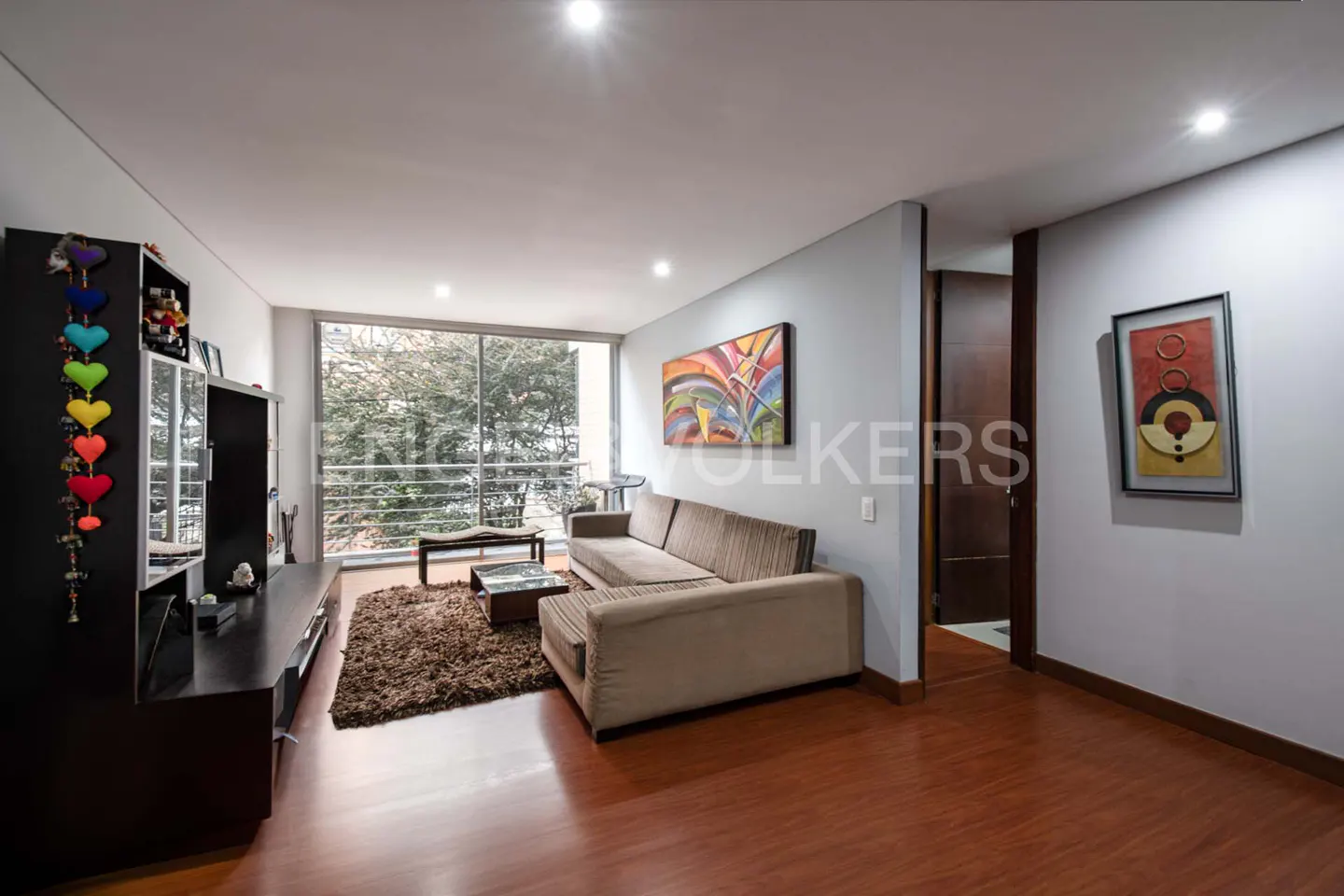Living room with brown sofa, shag rug, and black shelving unit with colorful heart decorations. Large window overlooks trees.