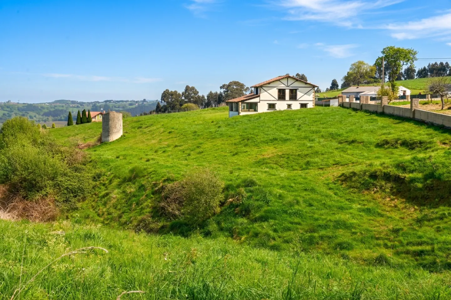 A white house with brown trim sits atop a lush green hill under a bright blue sky. A stone tower is visible in the distance.