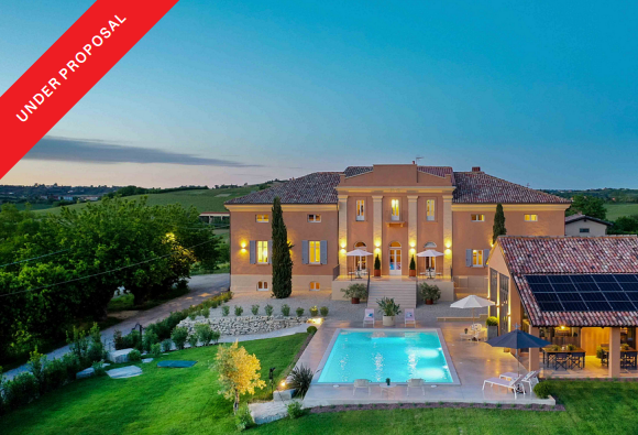 Large peach-colored villa at dusk with a glowing swimming pool, patio furniture, and a solar-paneled side building.