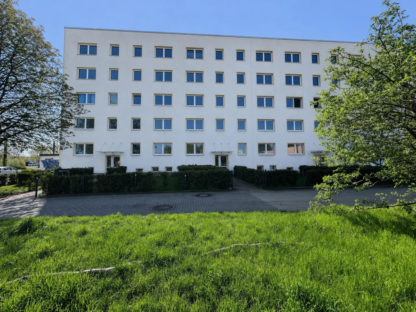 Exterior of a white, four-story apartment building with blue-framed windows, green lawn, and trees.