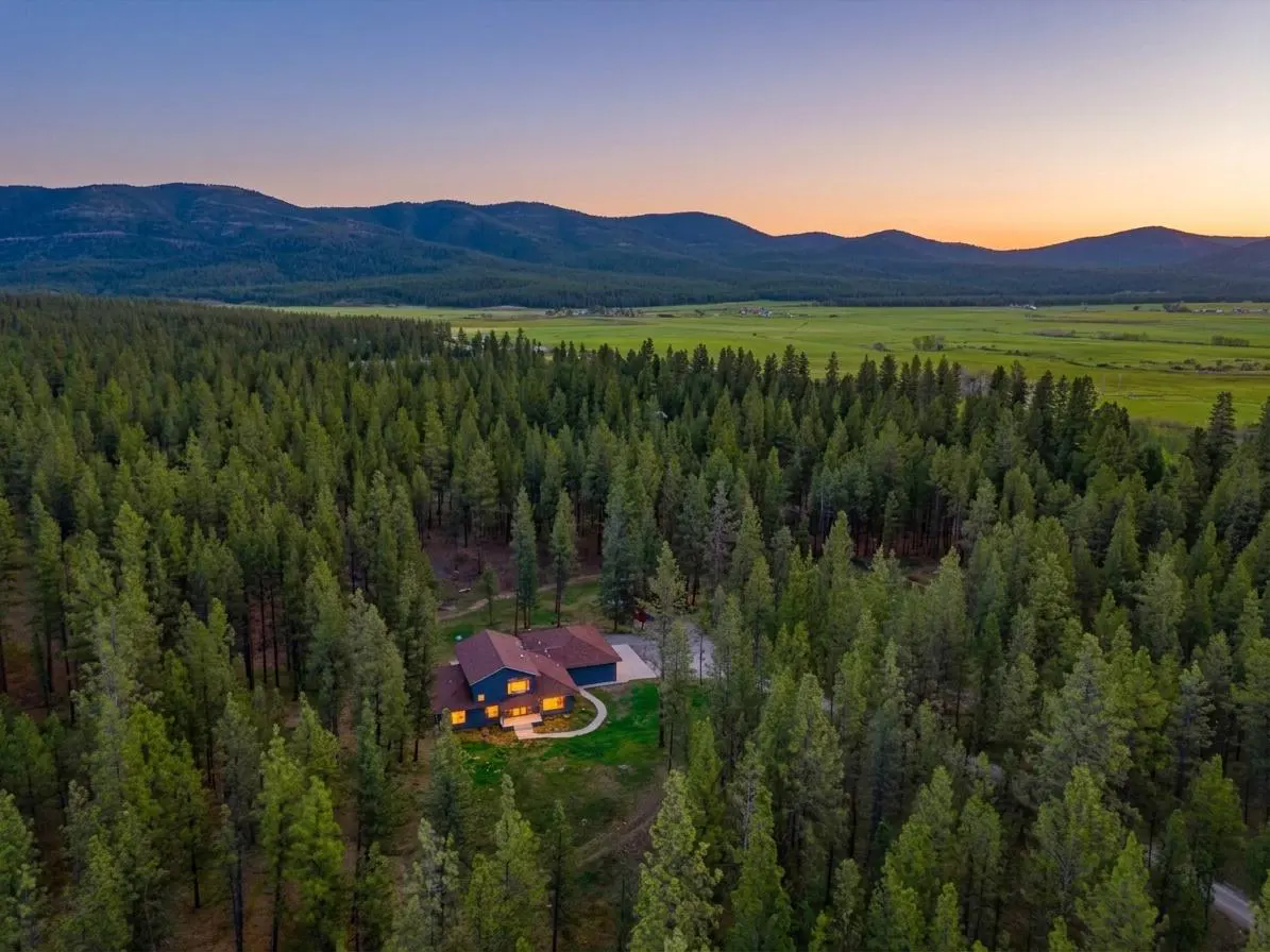 Aerial view of a blue house with a red roof nestled in a dense green forest, with mountains and a sunset sky in the background.