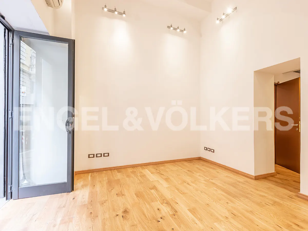 Bright, empty room with light wood floors, white walls, and an open glass door. Spotlights illuminate the space. Engel & Völkers logo on the wall.
