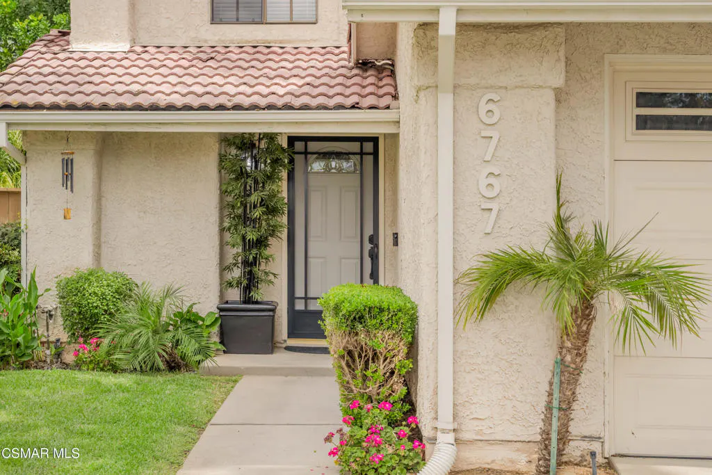 Beige house exterior with a red tile roof, white door, and address number 6767. A palm tree and green bushes decorate the front.