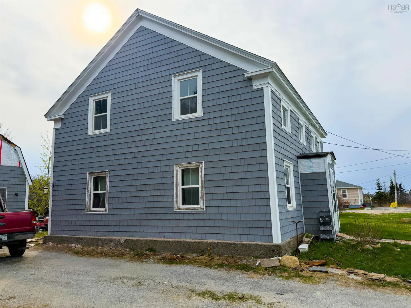 Two-story house with gray shingle siding and white trim, under a bright sun. A red pickup truck is parked nearby.