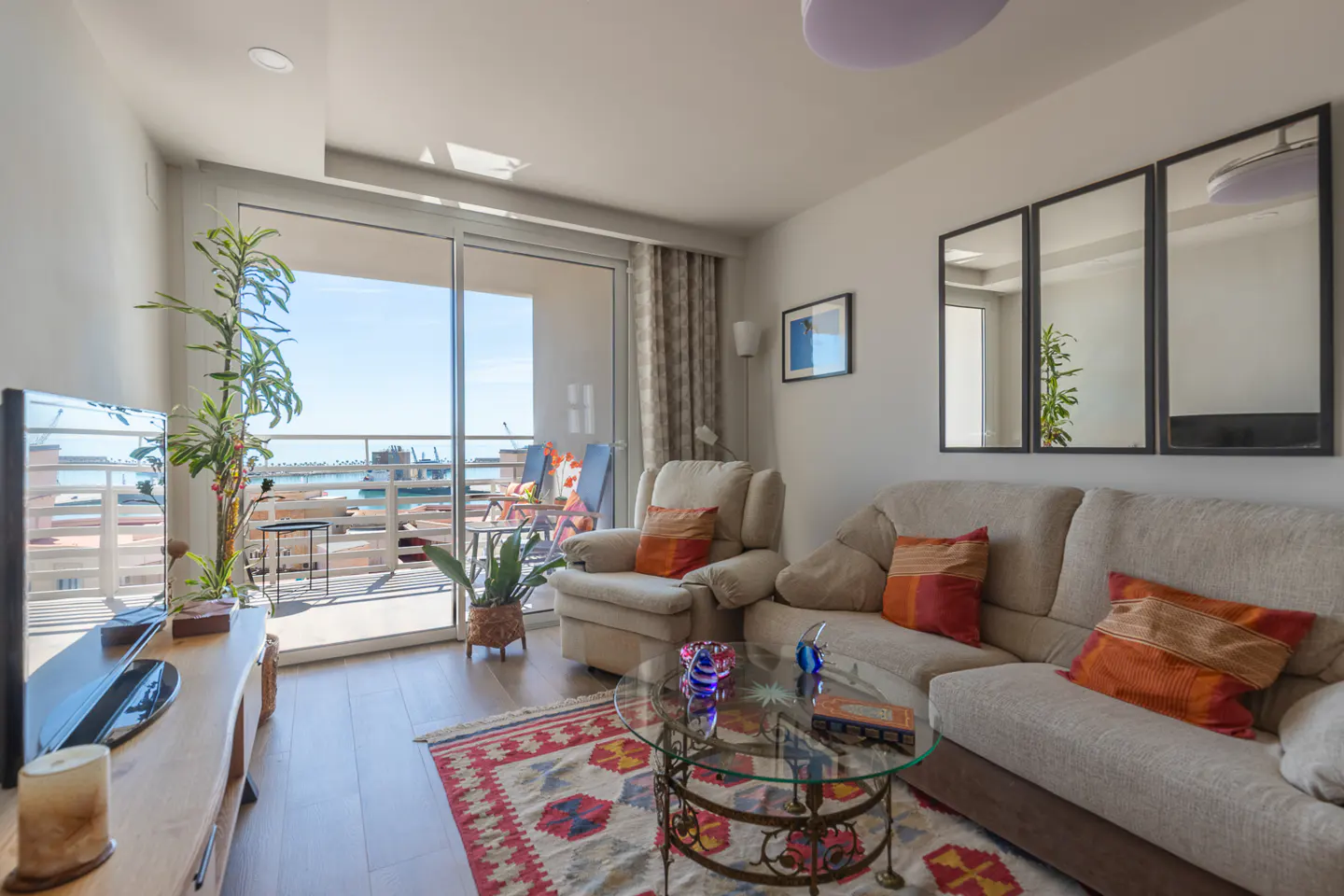Bright living room with a beige sofa, armchair, and glass table on a patterned rug. Balcony with ocean view visible through sliding glass doors.