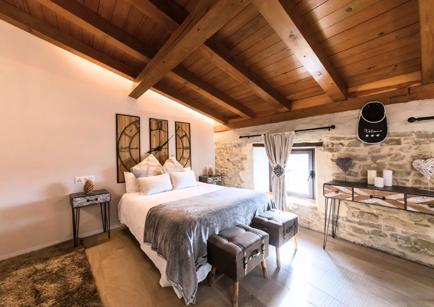 Bedroom with wooden ceiling beams, stone wall, and white linens. Three clock faces hang above the bed. Two brown ottomans sit at the foot of the bed.
