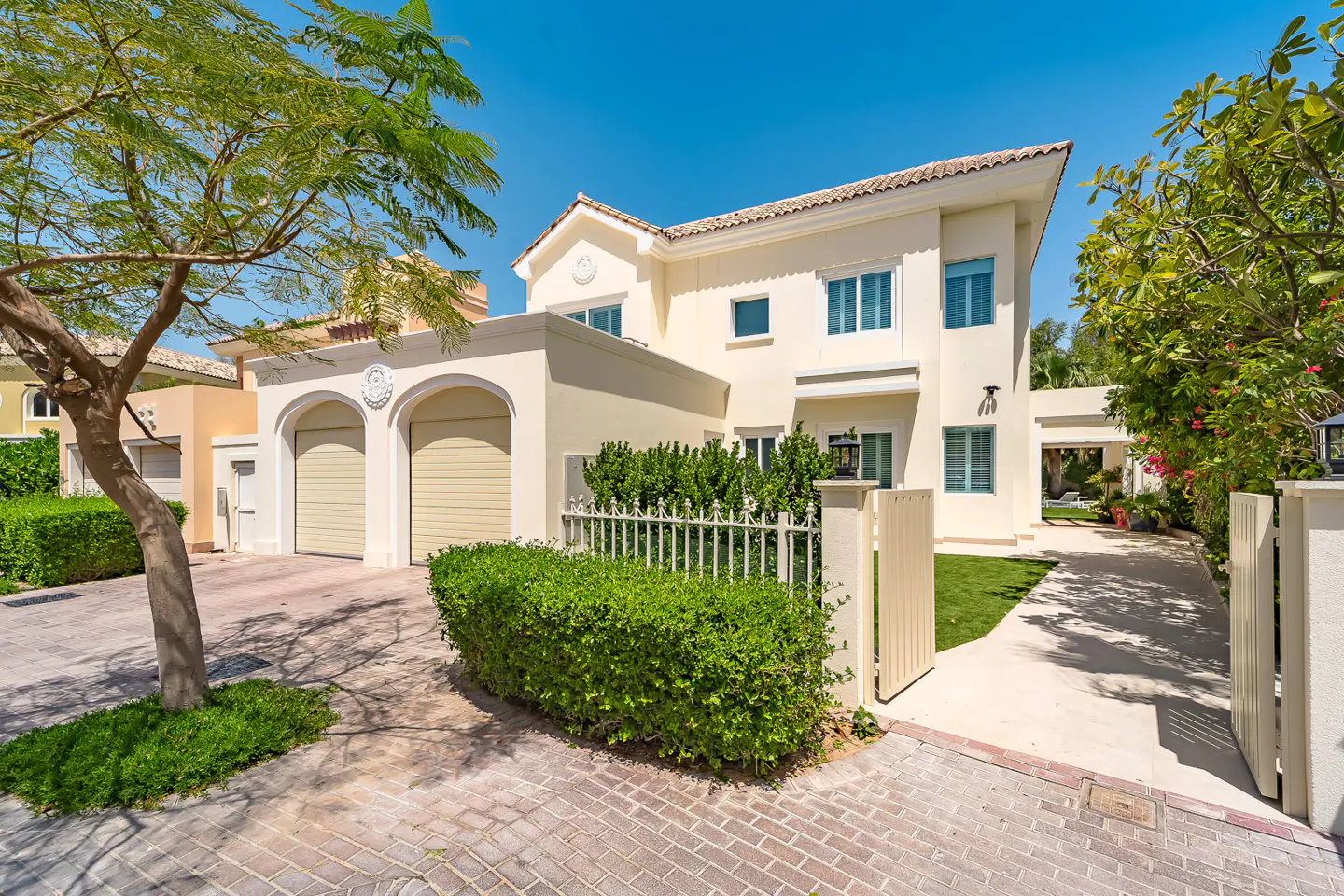 Two-story beige house with a double garage, green bushes, and a tree on a sunny day.