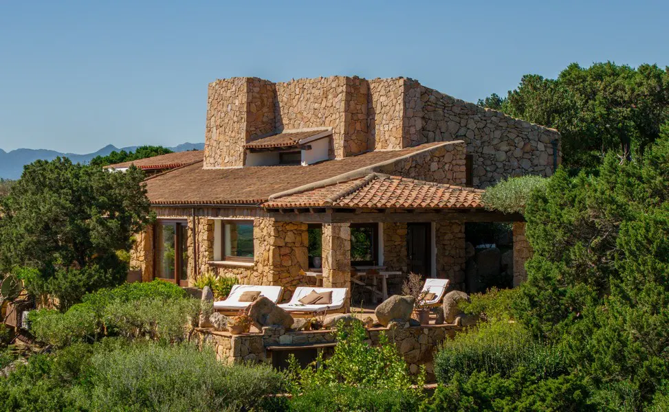 Stone house with a red tile roof, surrounded by green trees and bushes, with lounge chairs on a stone patio.
