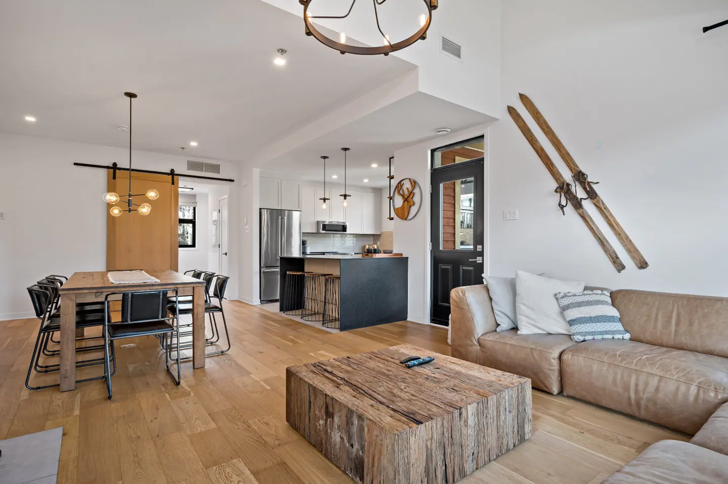 Open-concept living space with wood floors, dining table, leather sofa, and kitchen island. Skis hang on the wall.