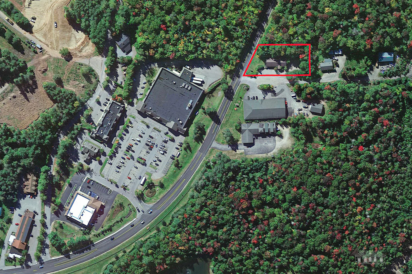 Aerial view of a commercial area with buildings, parking lots, and a road, surrounded by green and red trees. A red rectangle highlights a property.