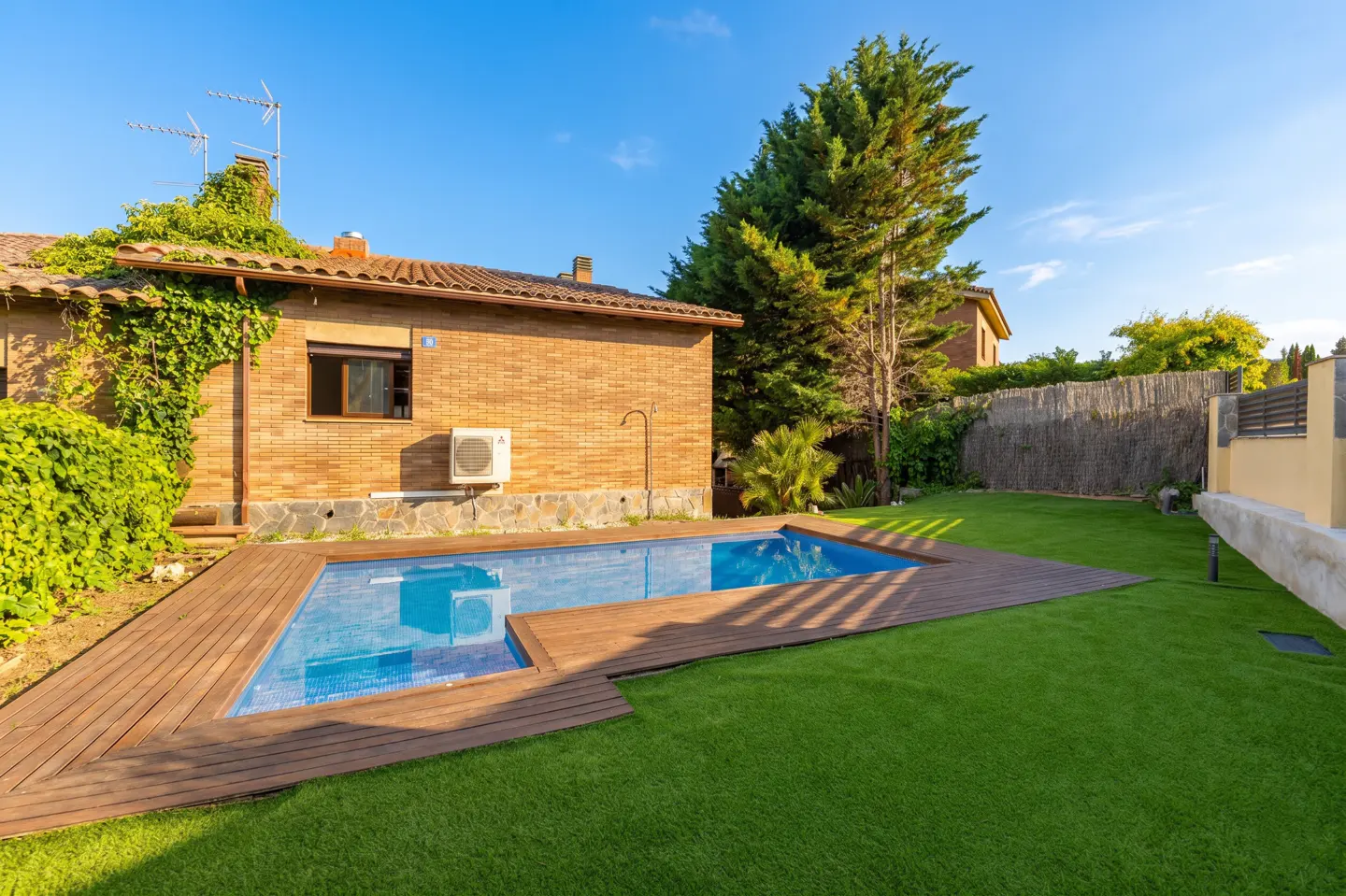 Backyard view of a brick house with a blue tiled pool, wooden deck, and green lawn under a sunny sky.