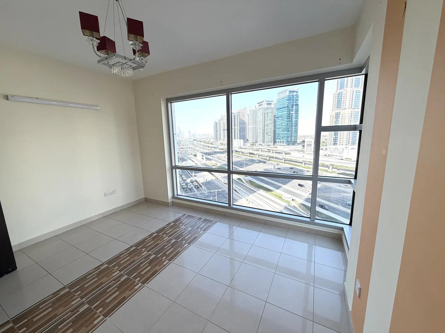 A bright, empty room with tile floors and a large window overlooking a city skyline with highways. A modern chandelier hangs from the ceiling.