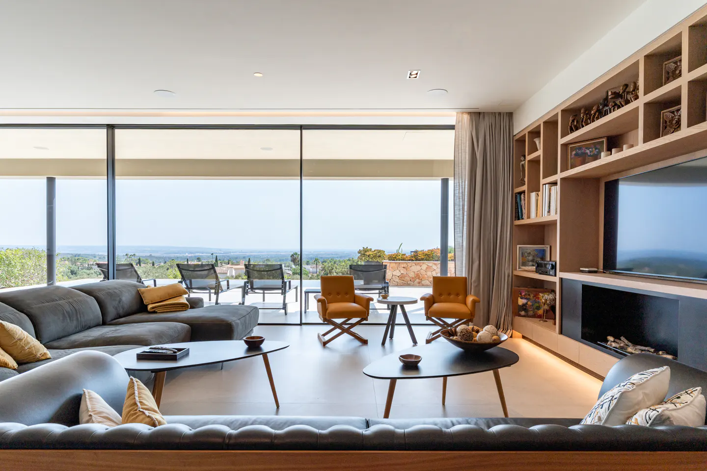 A modern living room with a large window view. Gray sofas, orange chairs, and black tables furnish the space. A built-in bookshelf surrounds a TV and fireplace.