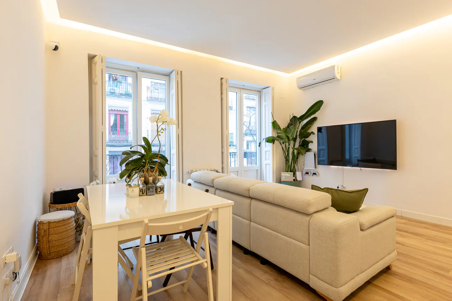 Bright living room with white walls, wood floors, and recessed lighting. A white table and beige sofa are visible. Windows overlook a city street.