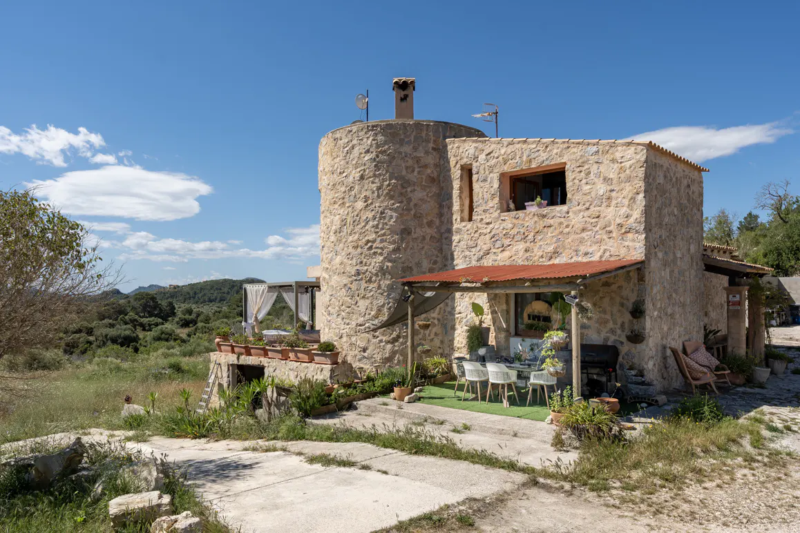 Stone house with a round tower under a blue sky. Outdoor dining area with white chairs and a red corrugated metal roof.