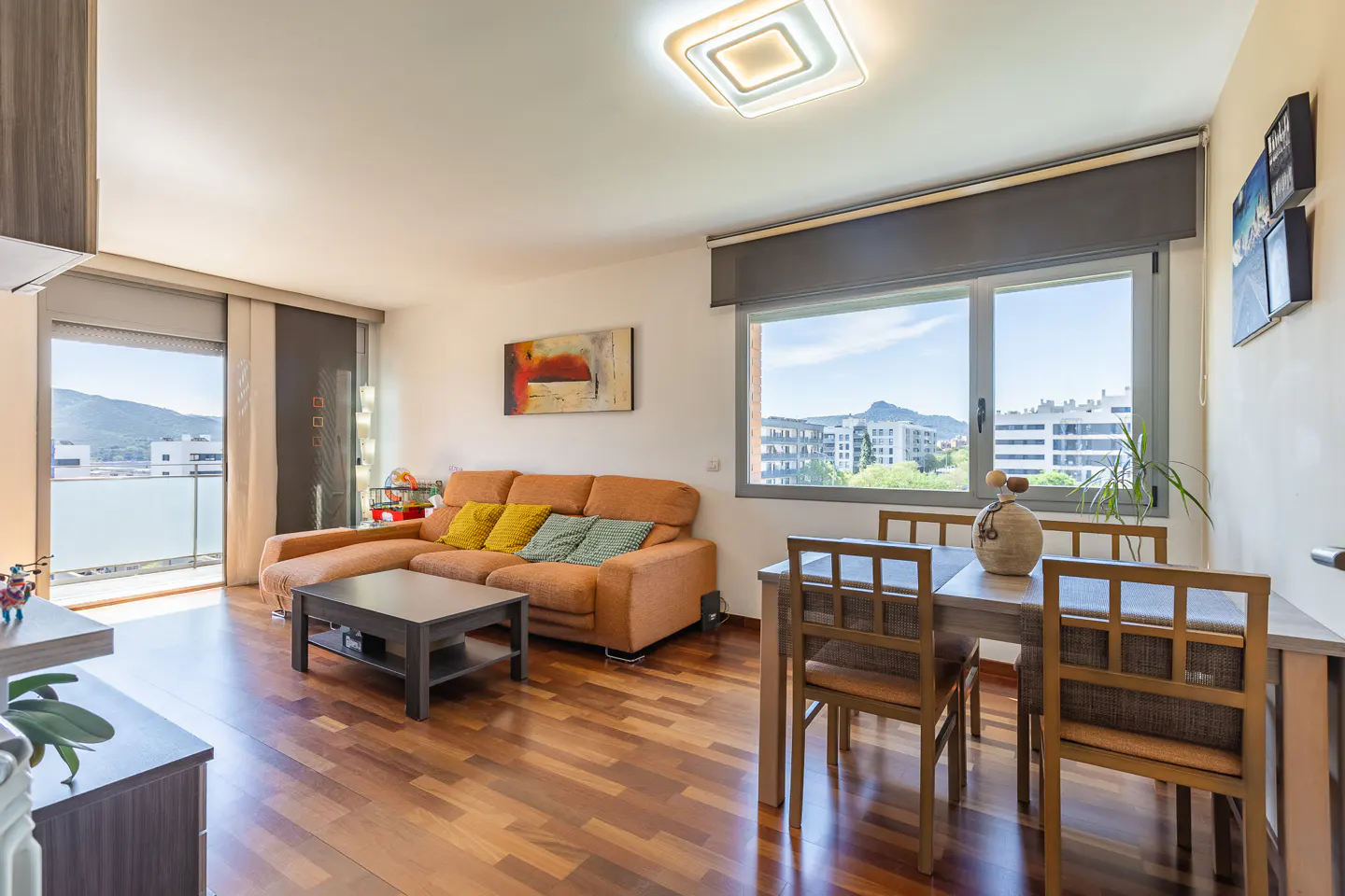 Bright living room with hardwood floors, orange sofa with yellow pillows, and a dining table set for four. Balcony view of mountains.