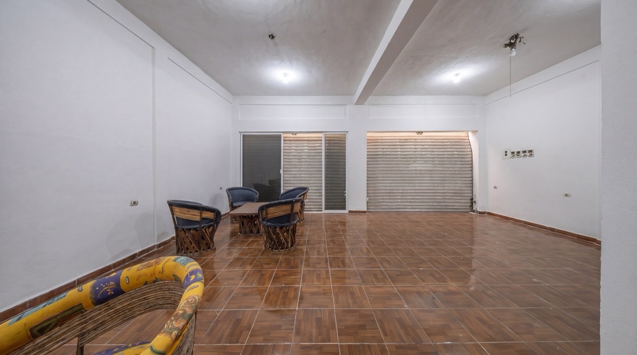 Bright, empty room with brown tile floor. Wicker chairs surround a small table. A closed metal shutter door is on the back wall.