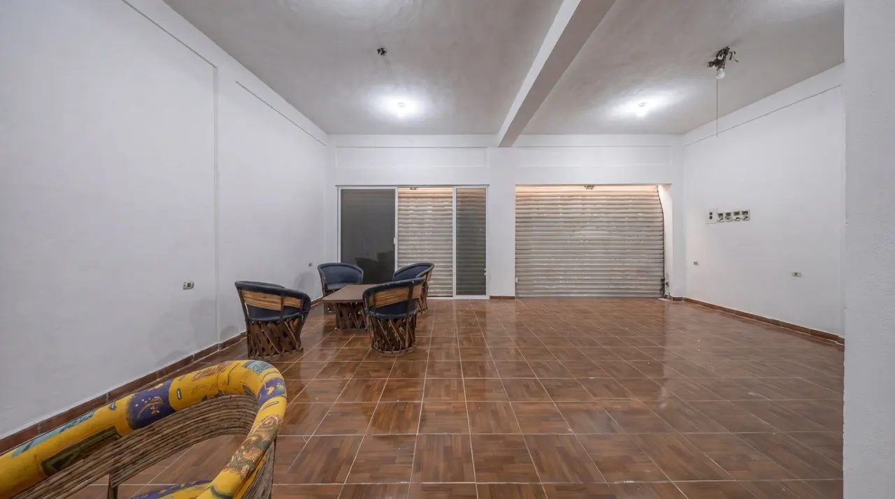 Bright, empty room with brown tile floor. Wicker chairs surround a small table. A closed metal shutter door is on the back wall.
