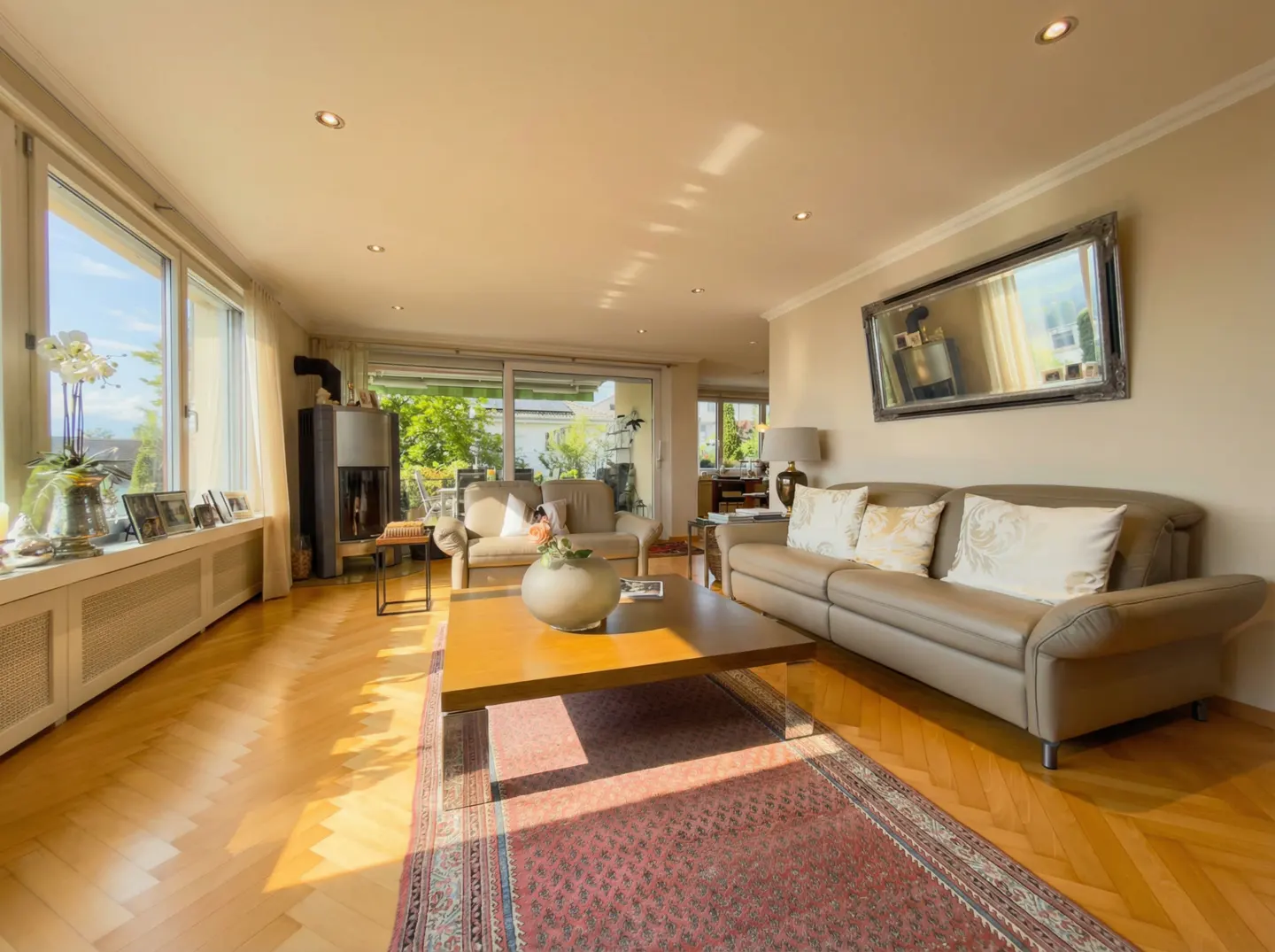 Bright living room with hardwood floors, a brown sofa, a wood coffee table, and a red patterned rug. Large windows offer natural light.