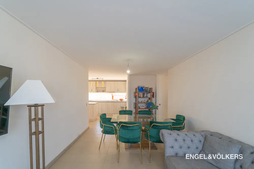 Living room with a glass table, green velvet chairs, and a gray sofa. Kitchen in the background. Engel & Völkers logo in the corner.