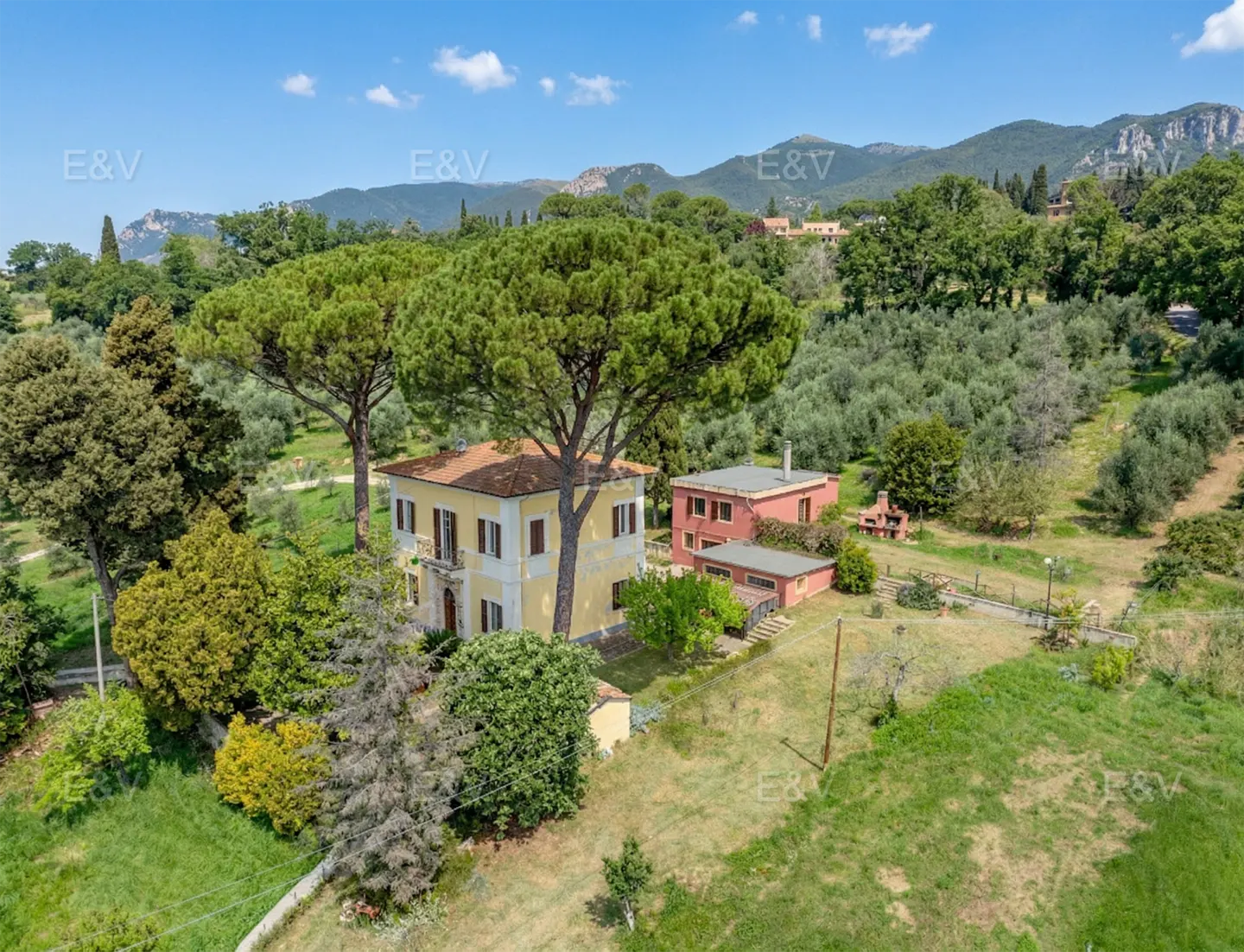 Aerial view of a yellow two-story house with brown shutters, surrounded by trees and a red outbuilding. Mountains are visible in the background.