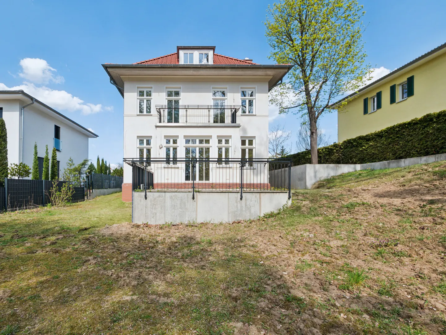 Two-story white house with a red roof, black railings, and a concrete foundation on a grassy hill.