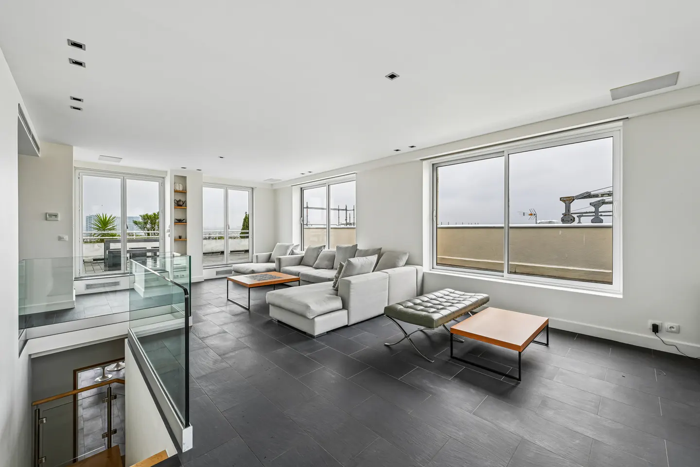 Bright, modern living room with gray sectional sofa, dark floors, and large windows overlooking a balcony.