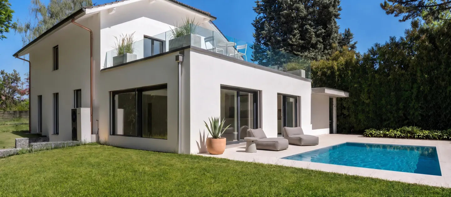 Modern white two-story house with a backyard pool, patio lounge chairs, and a glass-railed balcony under a clear blue sky.