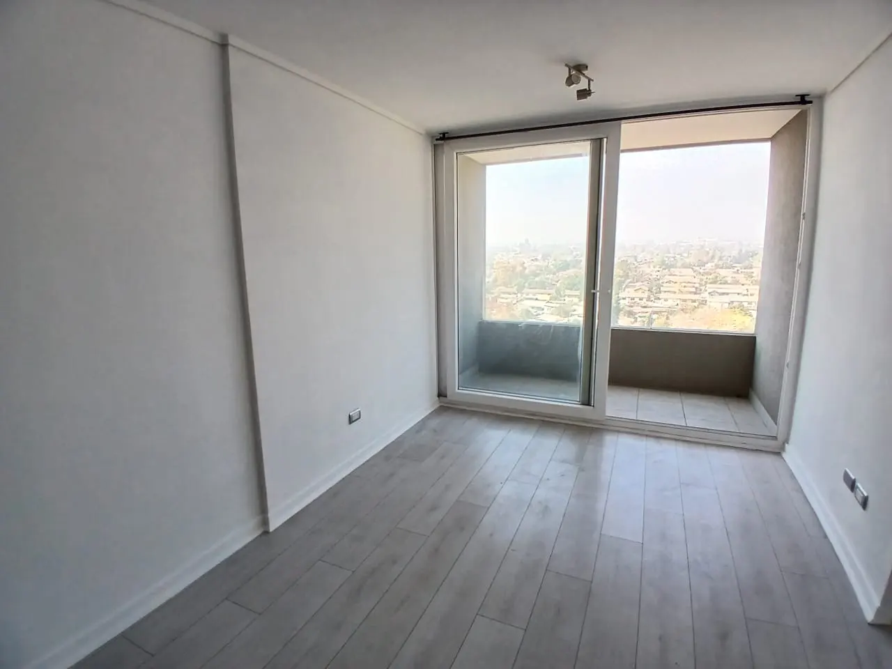 Bright, empty room with white walls, light wood floors, and sliding glass doors to a balcony overlooking a city.