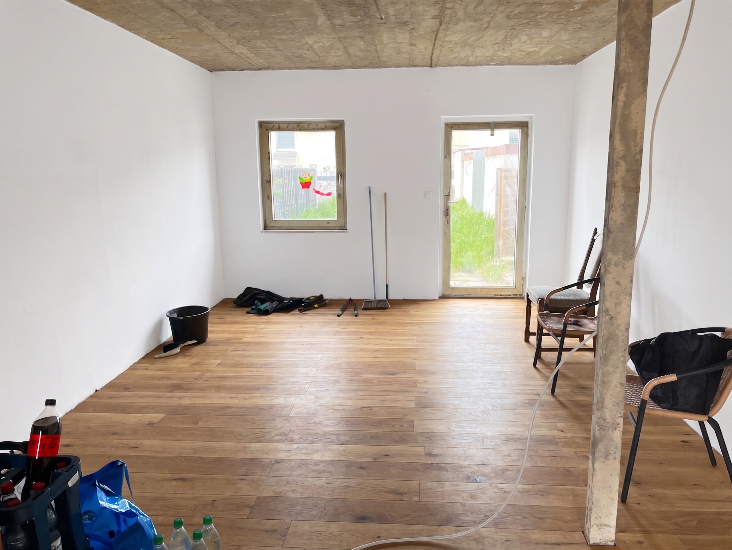 Unfinished room with white walls, wood flooring, a concrete ceiling, a window, a glass door, and scattered furniture. [AI-generated text] [AI-modified]