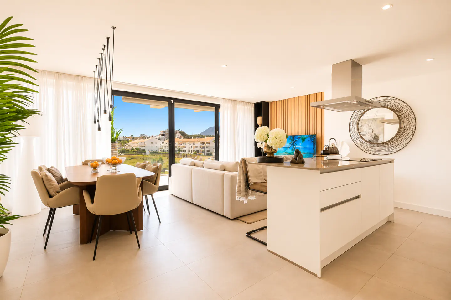 Bright, modern open-plan living space with dining table, white sofa, and kitchen island. Large windows offer a view of a sunny landscape.