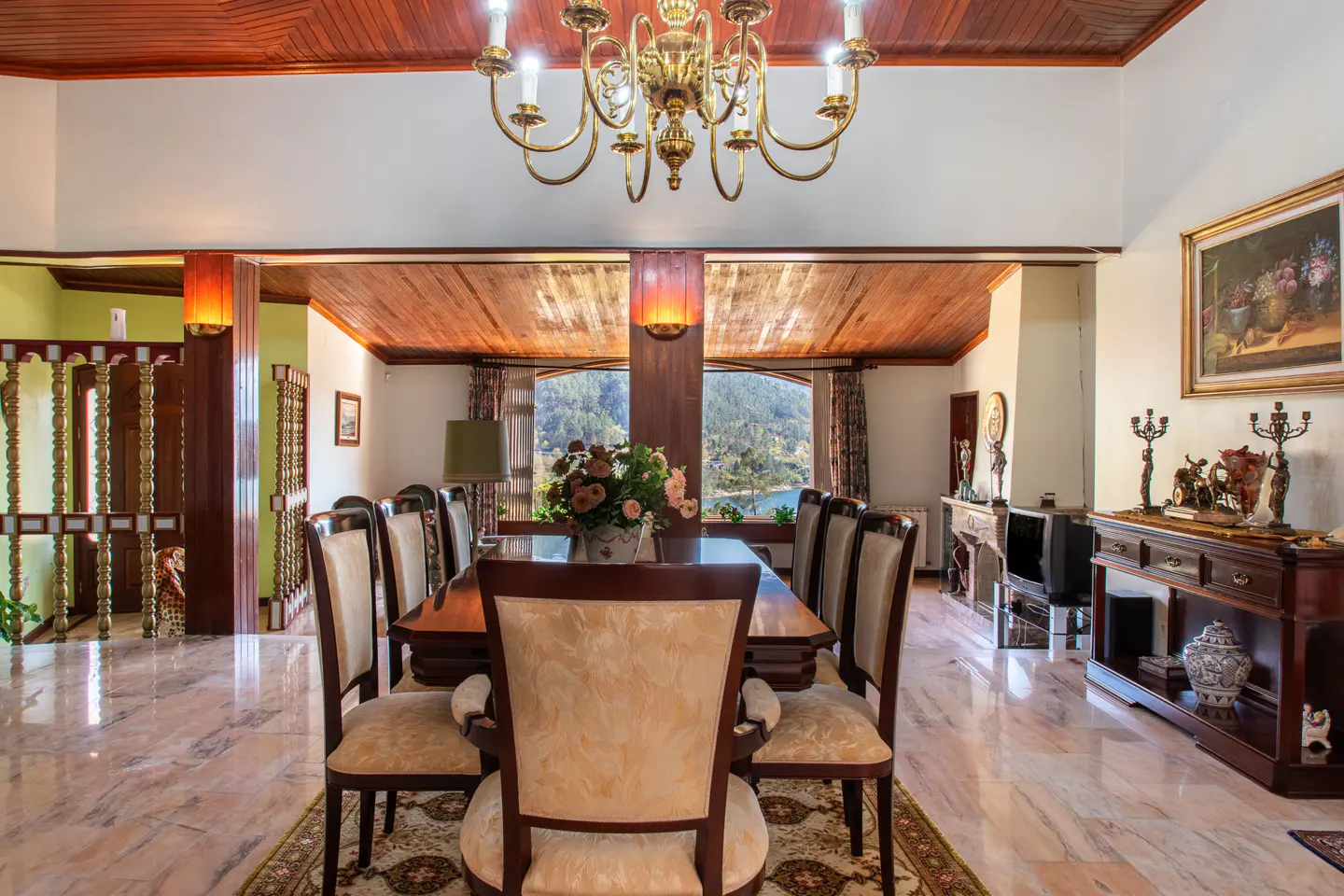 A dining room with a wood ceiling, marble floors, and a large table with upholstered chairs. A chandelier hangs above the table, and a window overlooks a lake.