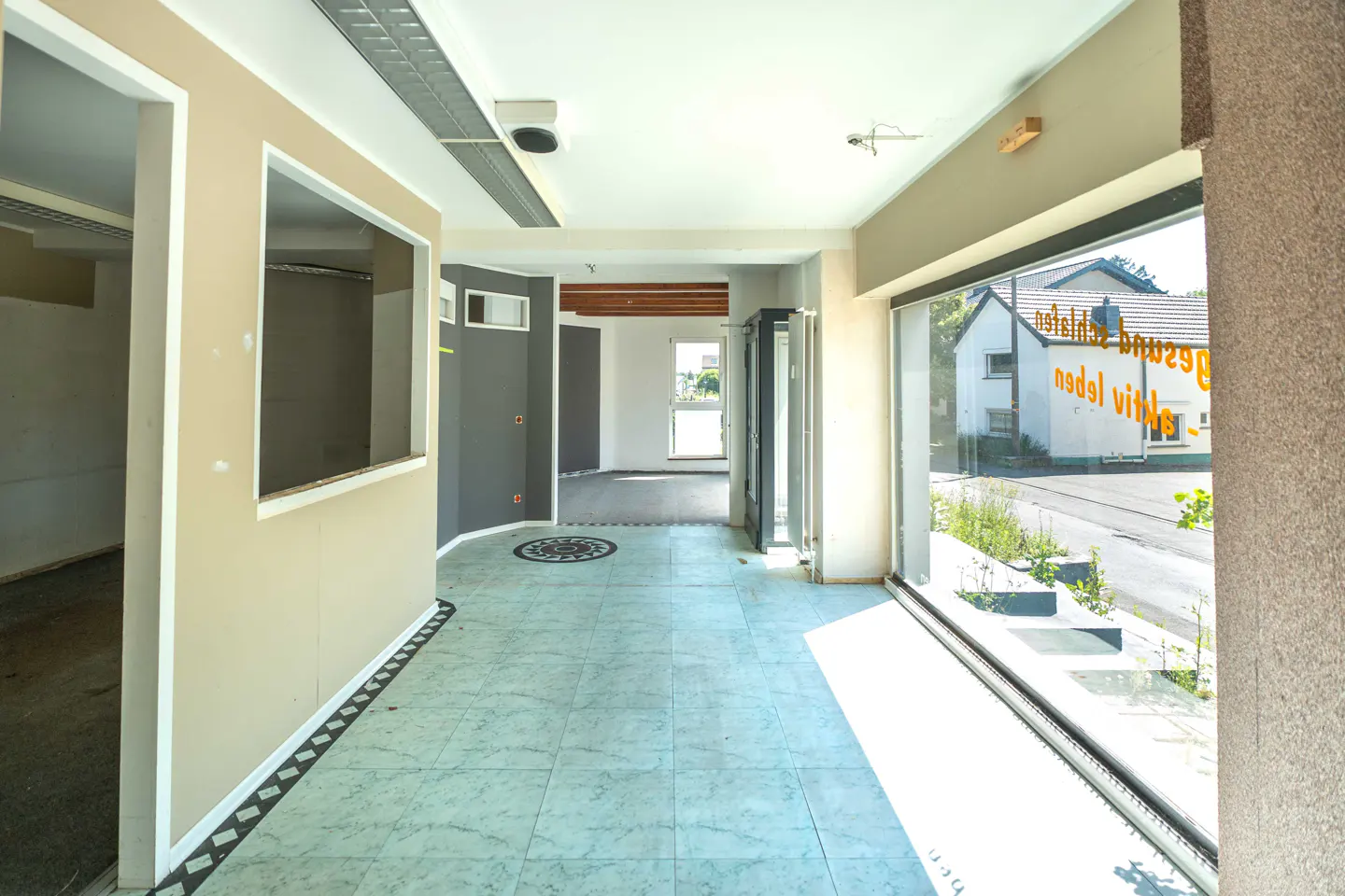 Interior view of a commercial space with light blue tile flooring, beige walls, and large windows. A building is visible outside.