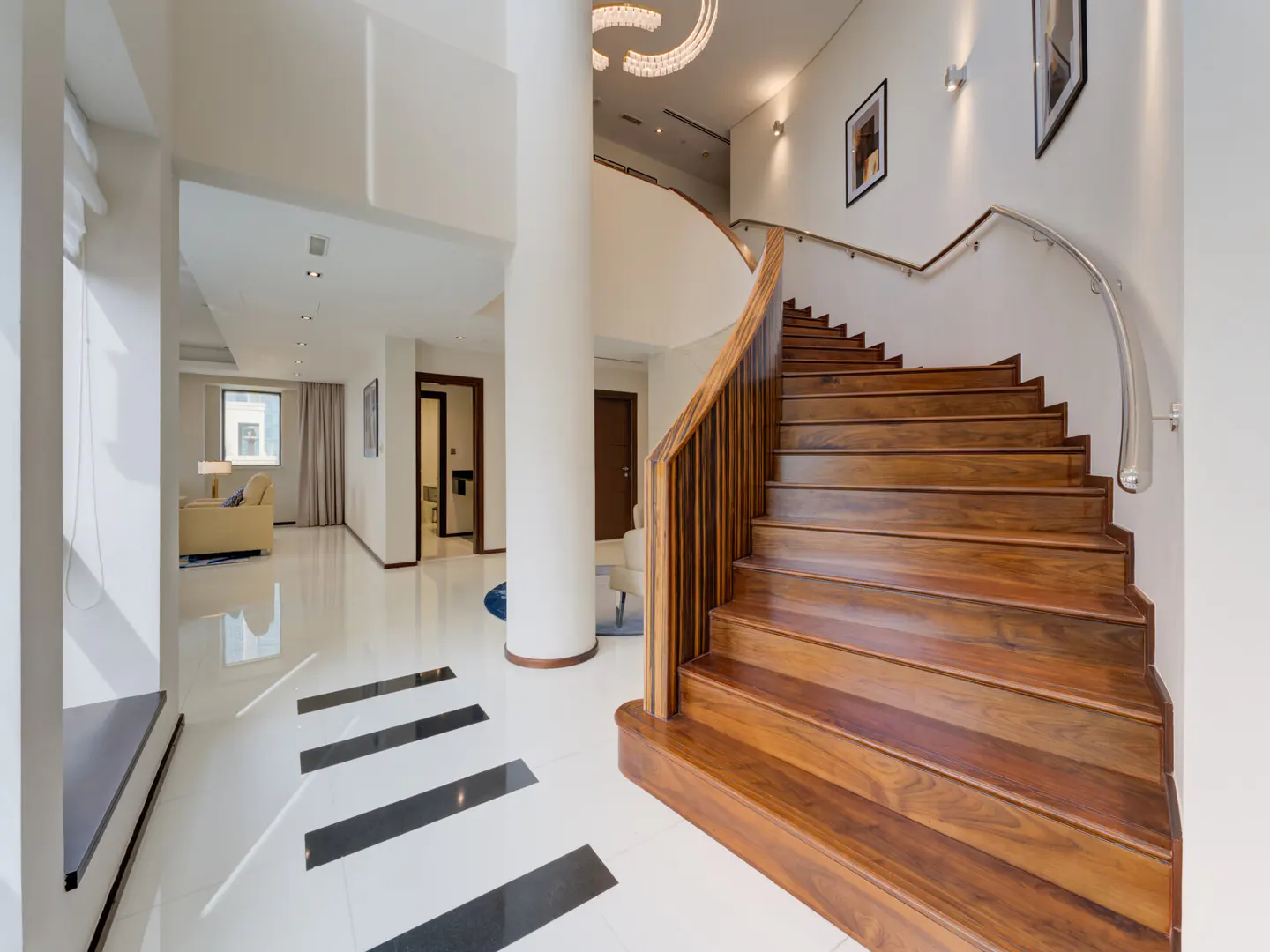 Bright foyer with a curved wood staircase, white walls, and a white tile floor with black accents. A white column supports the upper floor.