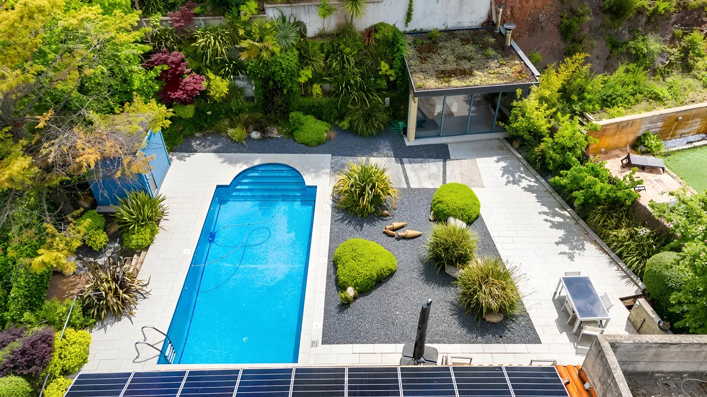 Aerial view of a backyard with a blue pool, lush green landscaping, a modern glass-walled structure with a green roof, and solar panels.