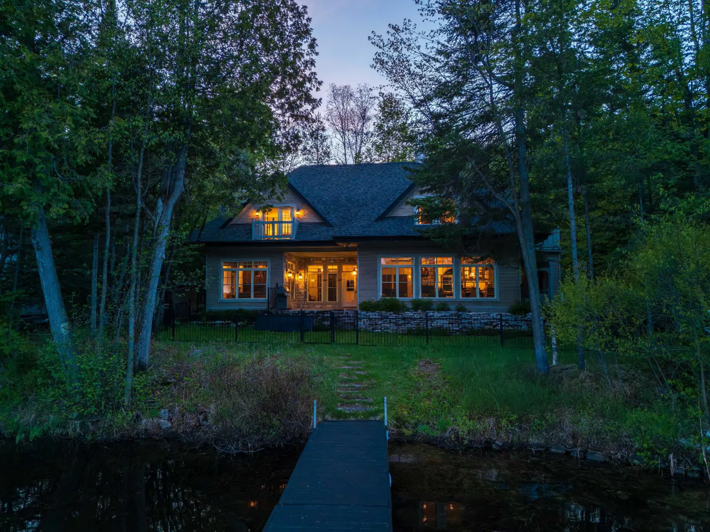 A lake house with a dock at dusk. The house is surrounded by trees and has lights on inside.