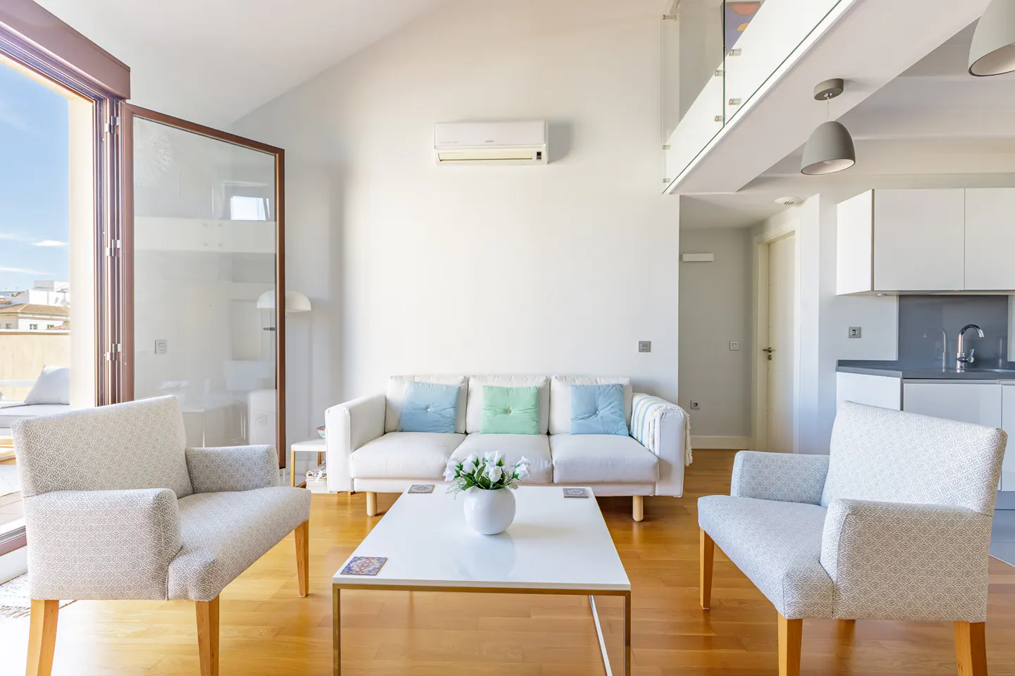 Bright living room with white sofa, blue and green pillows, two patterned armchairs, and a white coffee table with flowers. Balcony and kitchen visible.