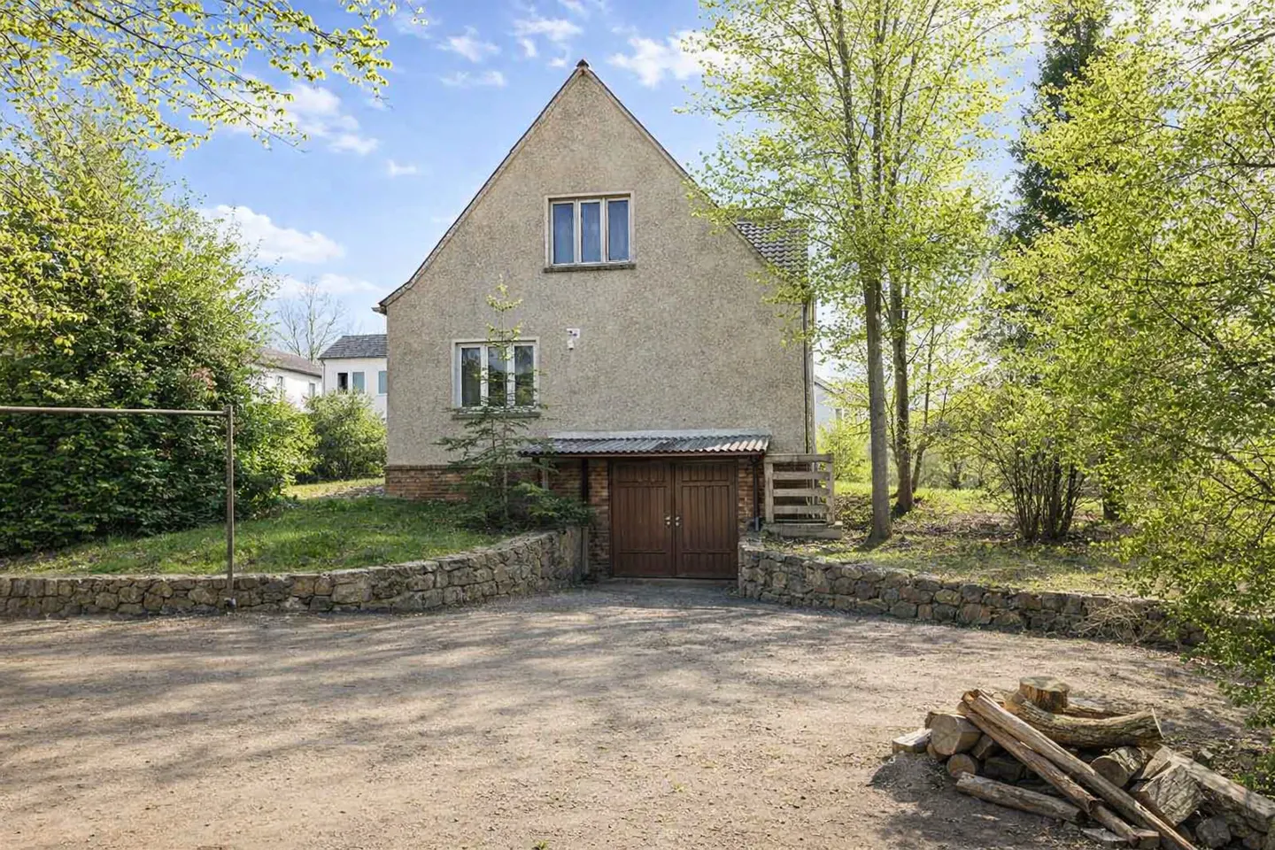 Two-story house with a brown garage door, stone wall, and dirt driveway. Trees surround the house.