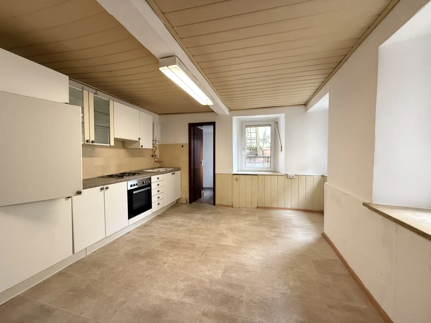 A bright, empty kitchen with white cabinets, a stove, and a window. The ceiling is wood paneled, and the floor is tiled.