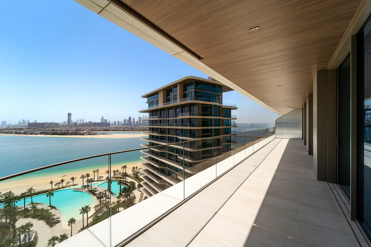 Balcony view of a modern high-rise building, pool, beach, and city skyline. Blue sky and water.